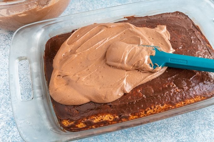 cake in a glass pan with a layer of pudding on top and a blue rubber spatula spreading chocolate frosting