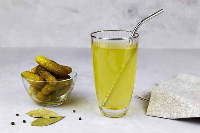 Cucumber pickle or pickle juice in glass with a metal tube for drinks , a bowl with pickled gherkins on light background. Trend drink, sports nutrition, healthy supplements.