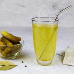 Cucumber pickle or pickle juice in glass with a metal tube for drinks , a bowl with pickled gherkins on light background. Trend drink, sports nutrition, healthy supplements.