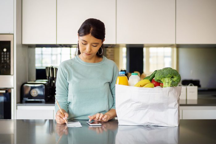 woman standing in her kitchen Checking her Grocery Budget; bag of groceries next to her on the counter