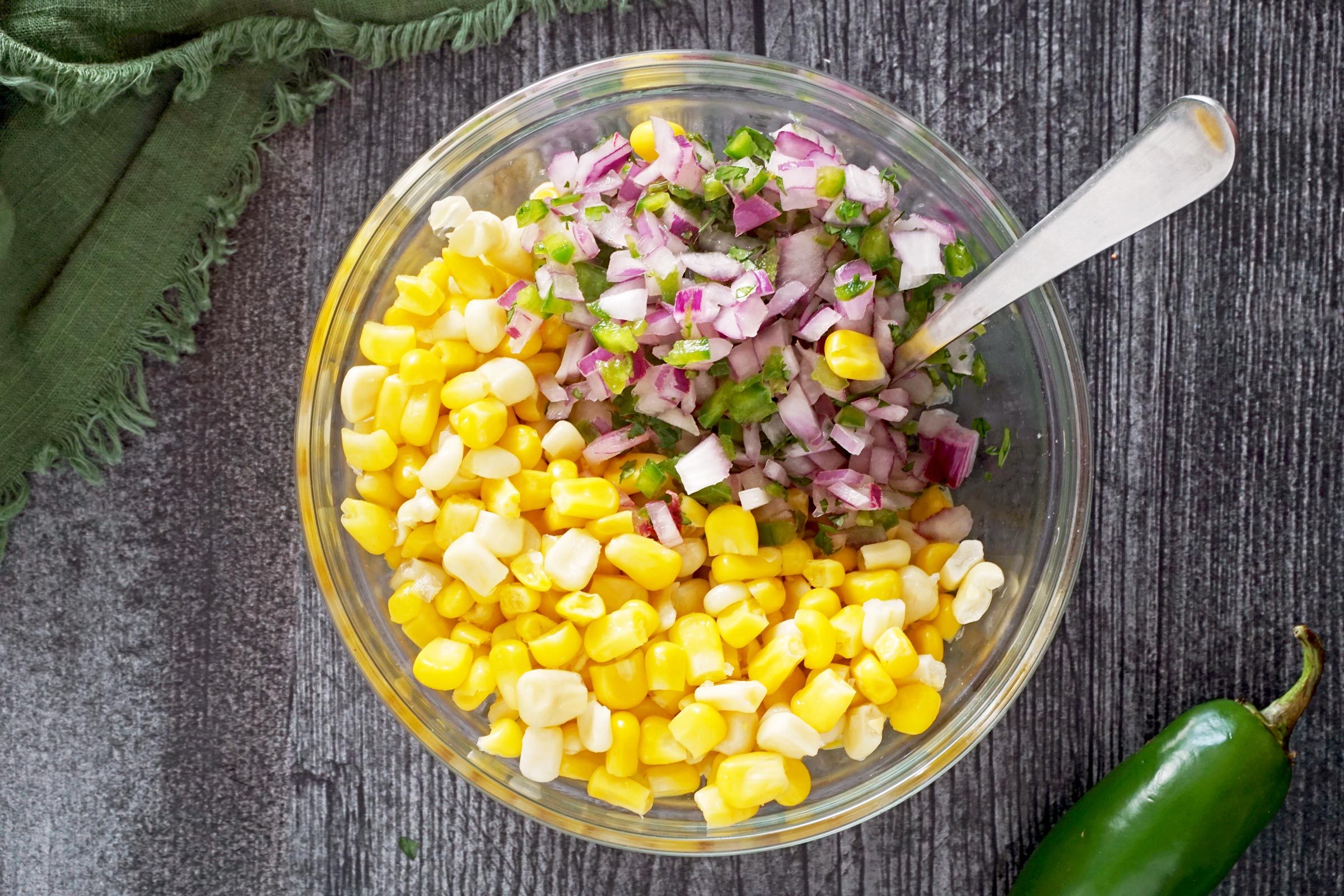 corn getting mixed with onions, peppers and herbs in a clear glass bowl