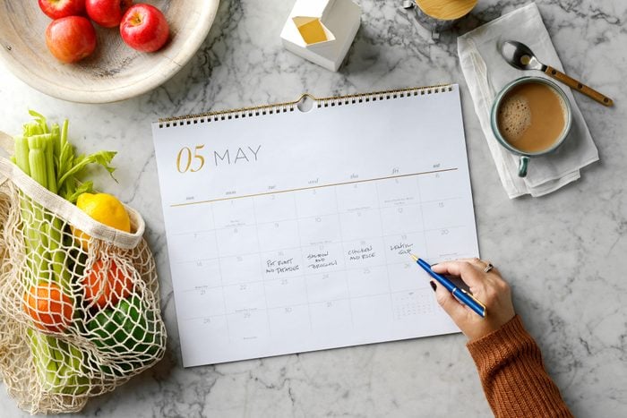 overhead view of hand writing meal plan ideas on a calendar laid on a marble counter. around the calendar are fresh groceries, fruits and vegetables, and a cup of coffee