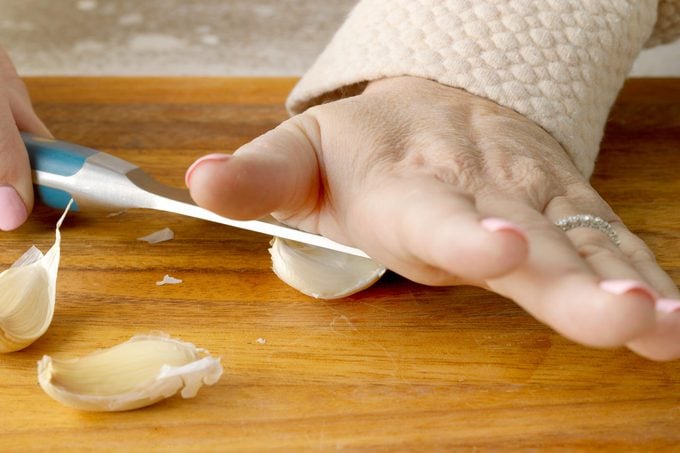 hands using a knife to press down on a clove of garlic on a wooden cutting board