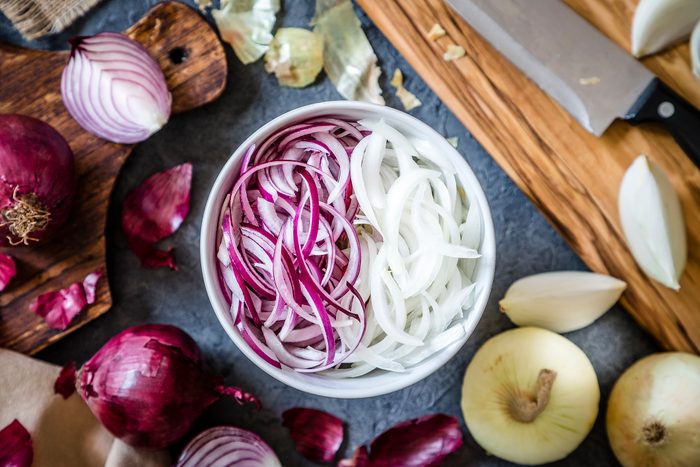 Top view of a bowl full of red and golden sliced onions surrounded by a wooden cutting board with a kitchen knife on top and whole and sliced golden and red onion