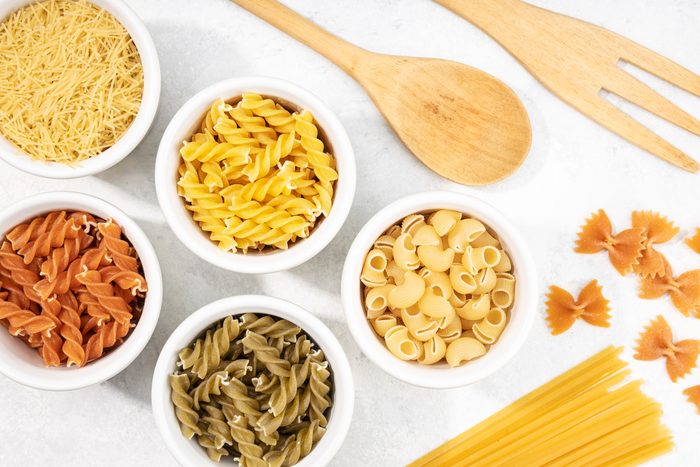 Top view of assorted uncooked pasta in small bowls with a wooden spoon and fork off the the side