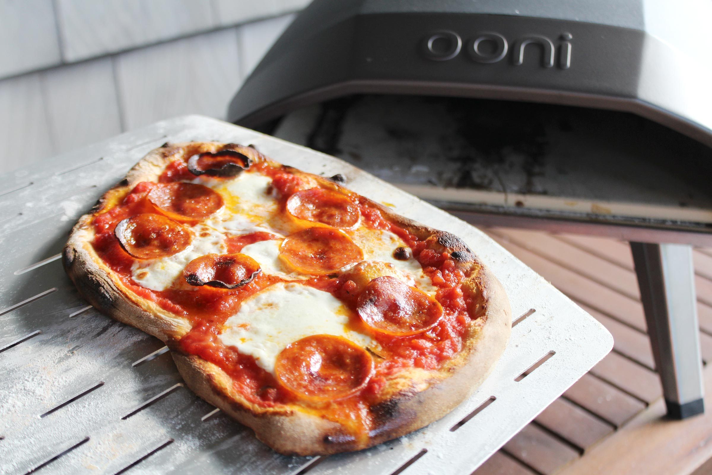 a freshly baked pizza on a pizza peel with oonie oven in the background