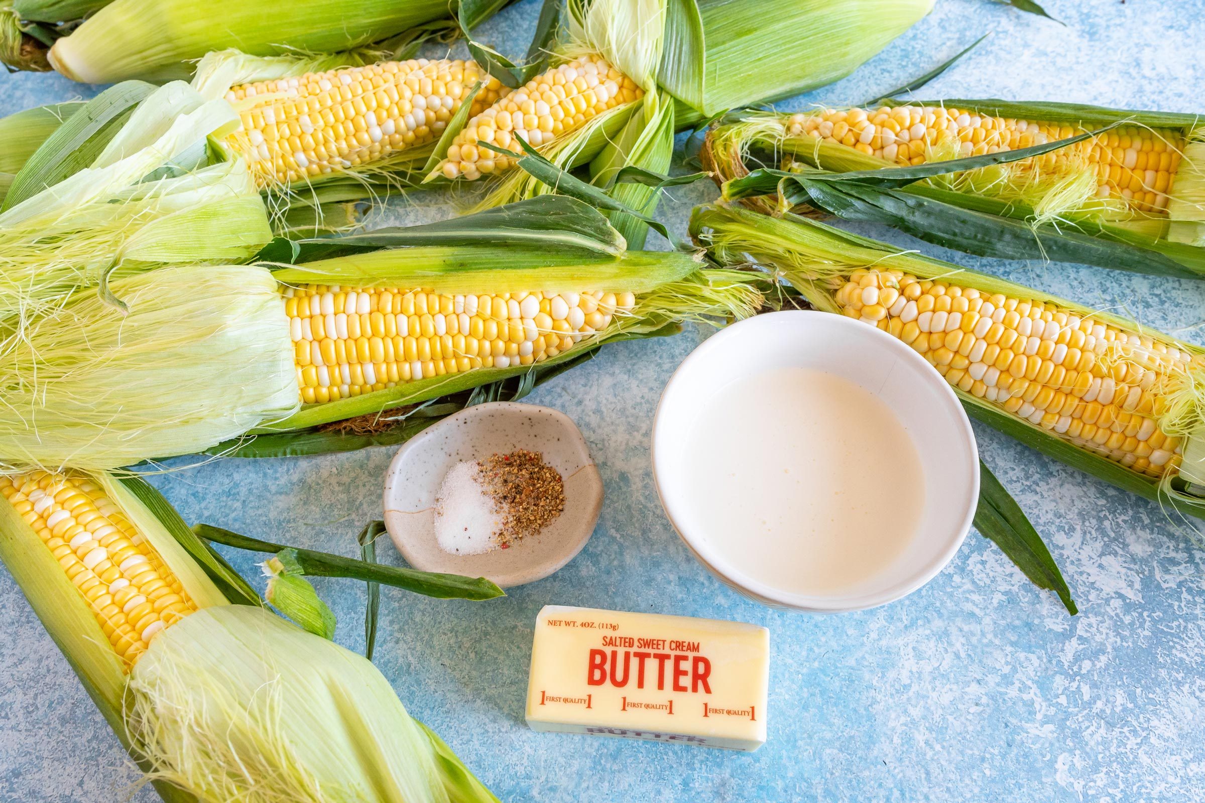 corn on the cob, butter, a dish of spices and a bowl of milk on a blue countertop