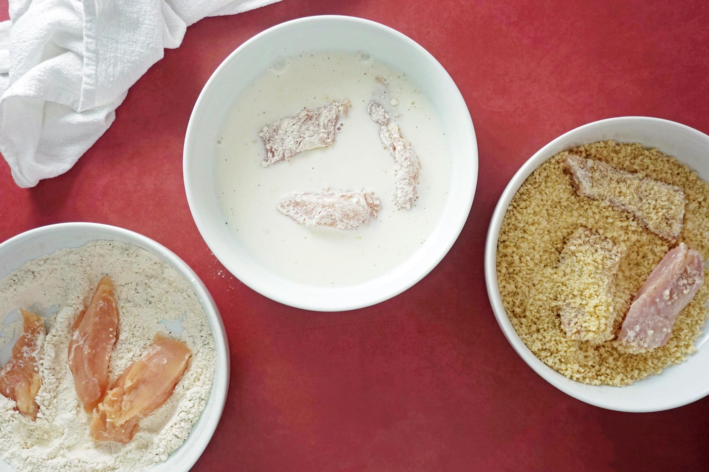 three small bowls with flour, milk and bread crumbs with small pieces of raw chicken in each demonstrating the steps for breading chicken
