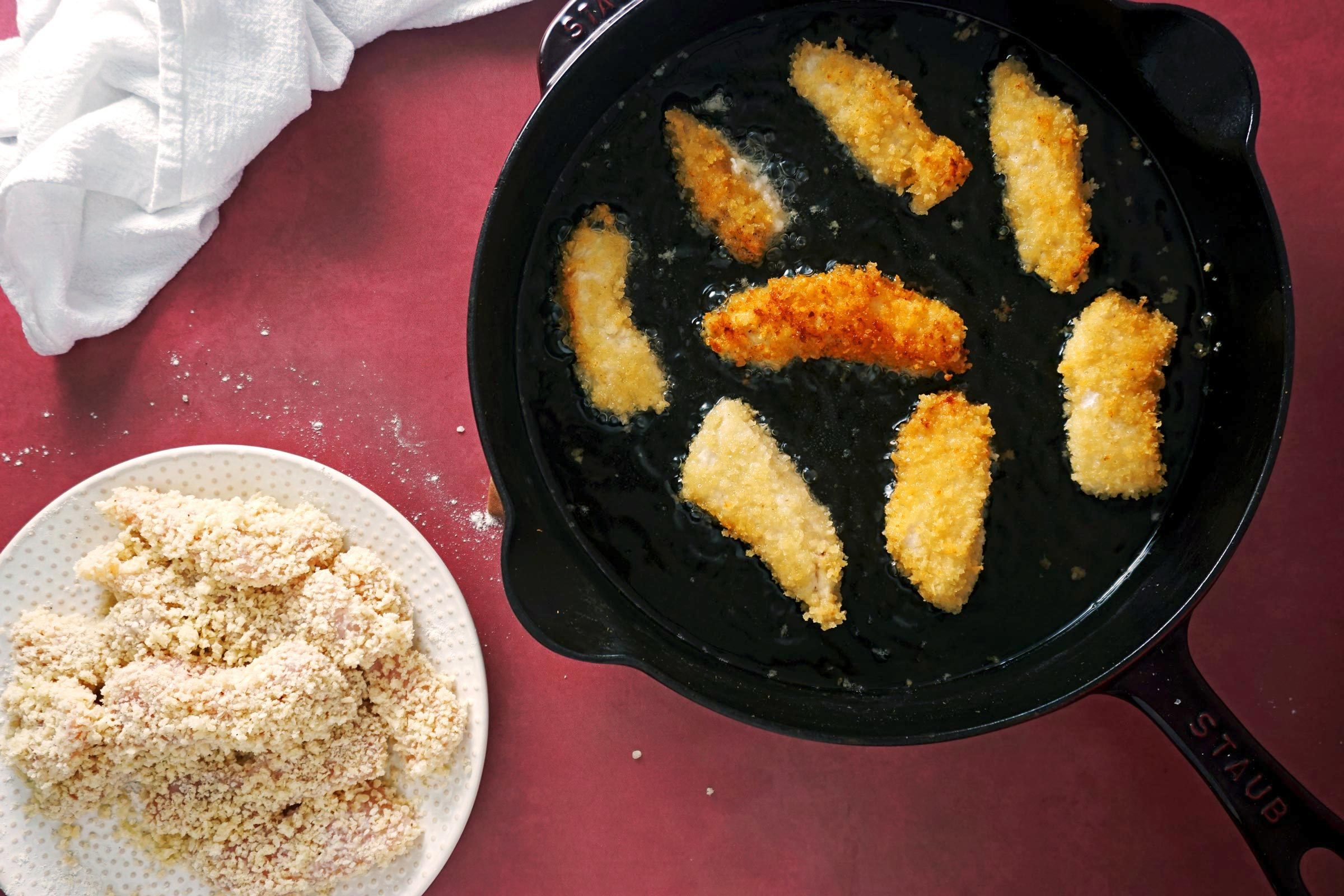 frying chicken in a cast iron pan with a plate of uncooked breaded chicken off to the side