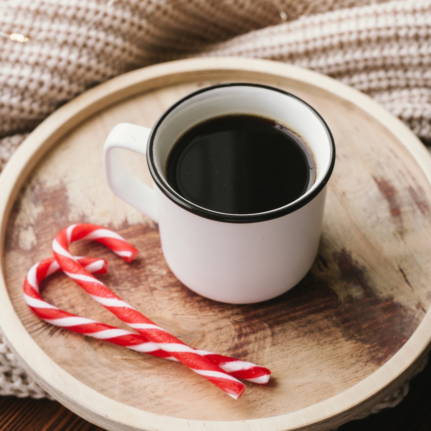White cup of coffee on wooden table, beige knitted fabric and red candy canes. Christmas cozy stil life.