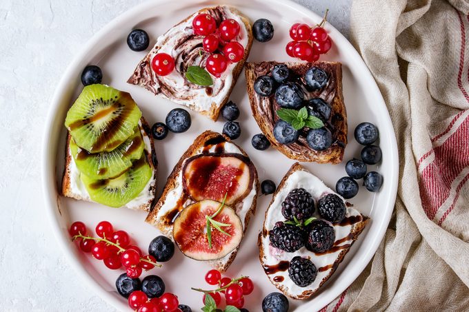 Variety of toasts with berries and cream cheese and chocolate swirl. Red currant, blueberries, sliced kiwi, figs in white plate over gray texture background.