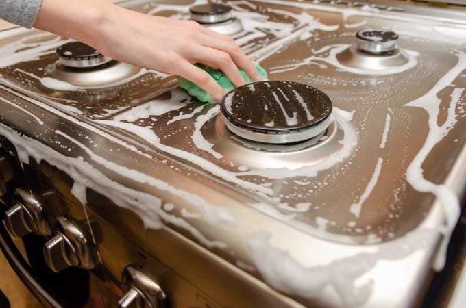 woman's hand washes a gray stainless steel gas stove with a green foam sponge. House cleaning