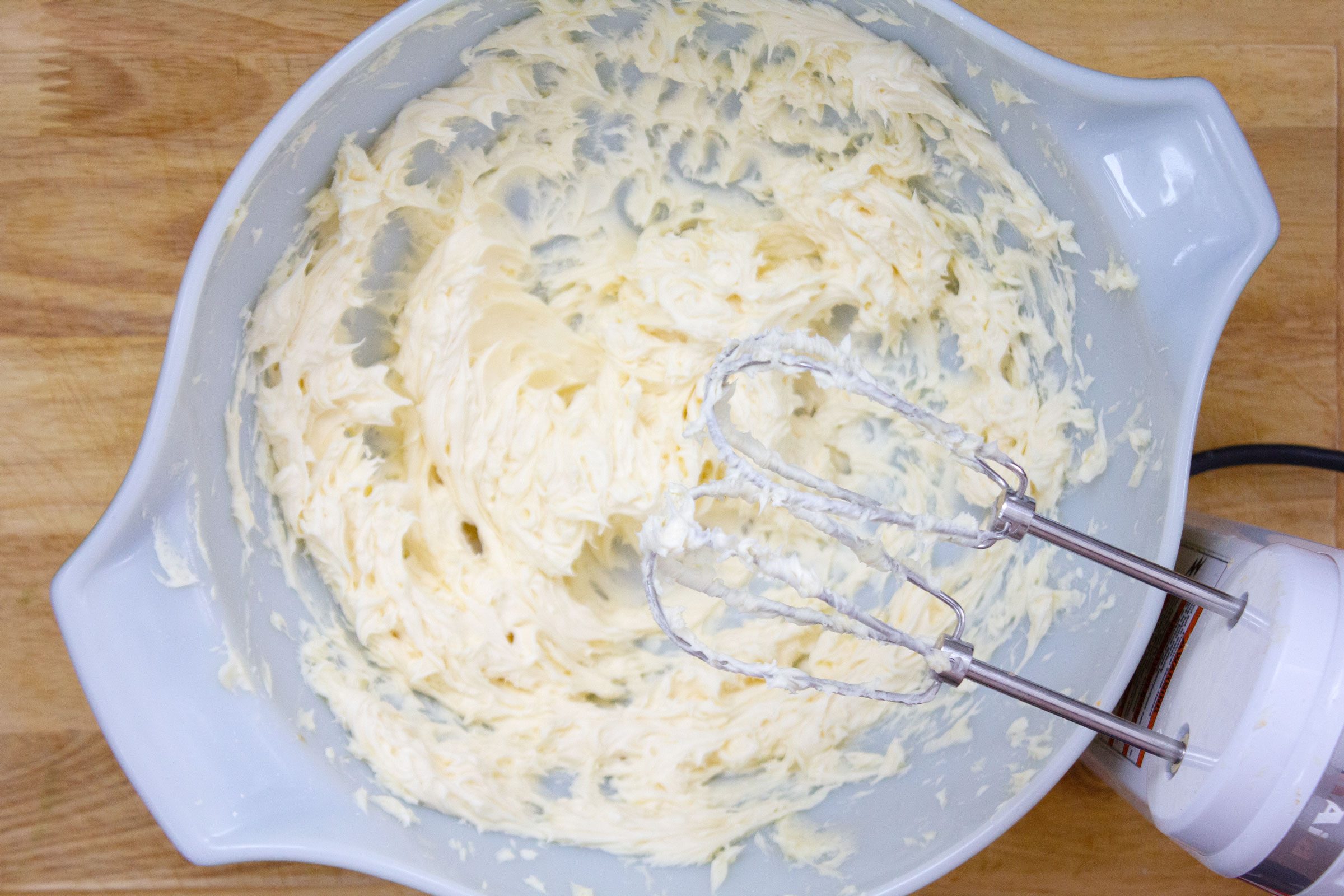 creaming butter in a plastic bowl on a wood surface with electric beaters resting on the side of the bowl