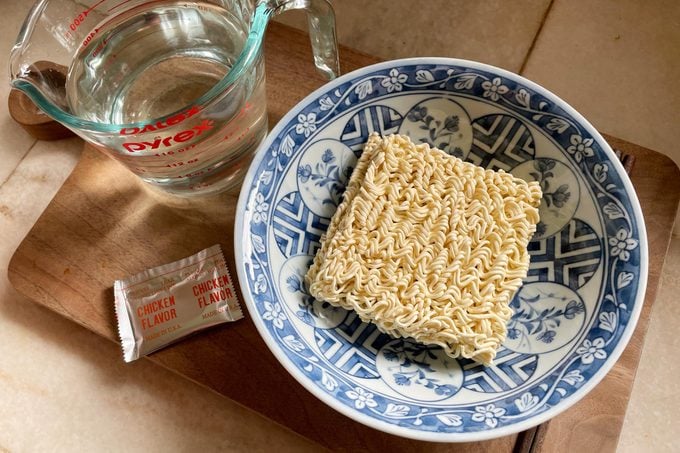 raw ramen noodles in a bowl, a measuring cup of water, and a flavor packet on a kitchen counter