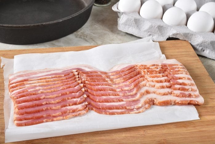 uncooked bacon slices on wax paper on a cutting board; eggs and pan in the background