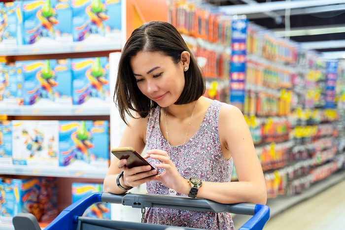 Woman checking shopping list by mobile phone, pulling shopping trolley at a supermarket