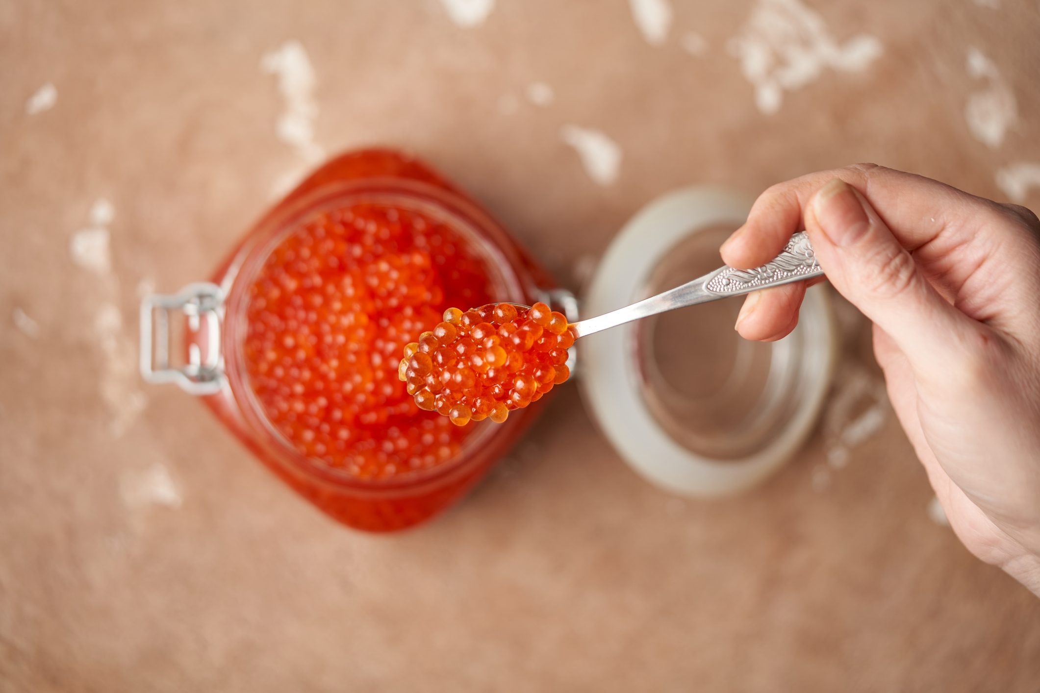 Female hand with a spoon with red caviar over an open glass jar filled with red caviar to the top on a beige textured stone surface. Seafood, delicacies, healthy nutrition. Place for text. Top view, shallow depth of field