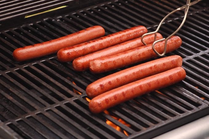 Six hot dogs cooking on a gas-fired barbeque grill. Flames licking the bottoms of the polish dogs are visible below the grill's grates