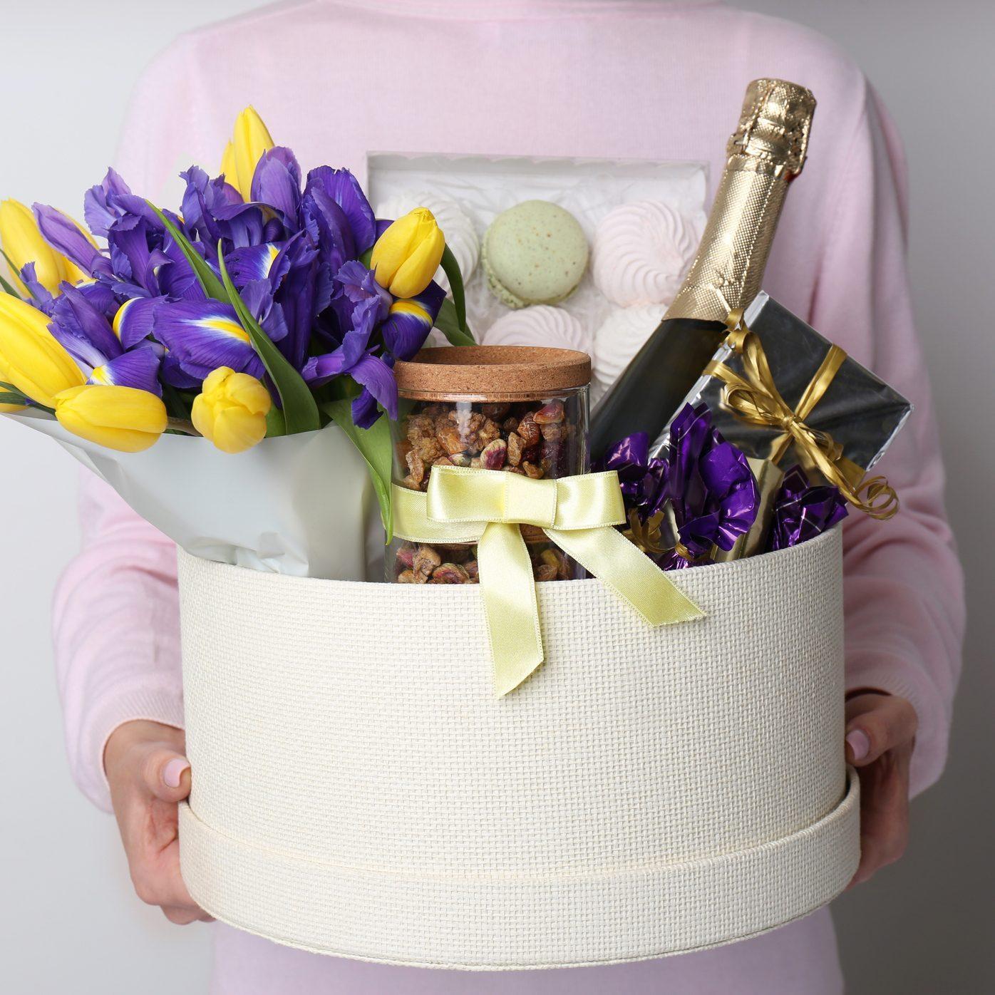 Woman holding basket with gifts, bouquet and champagne on light grey background, closeup