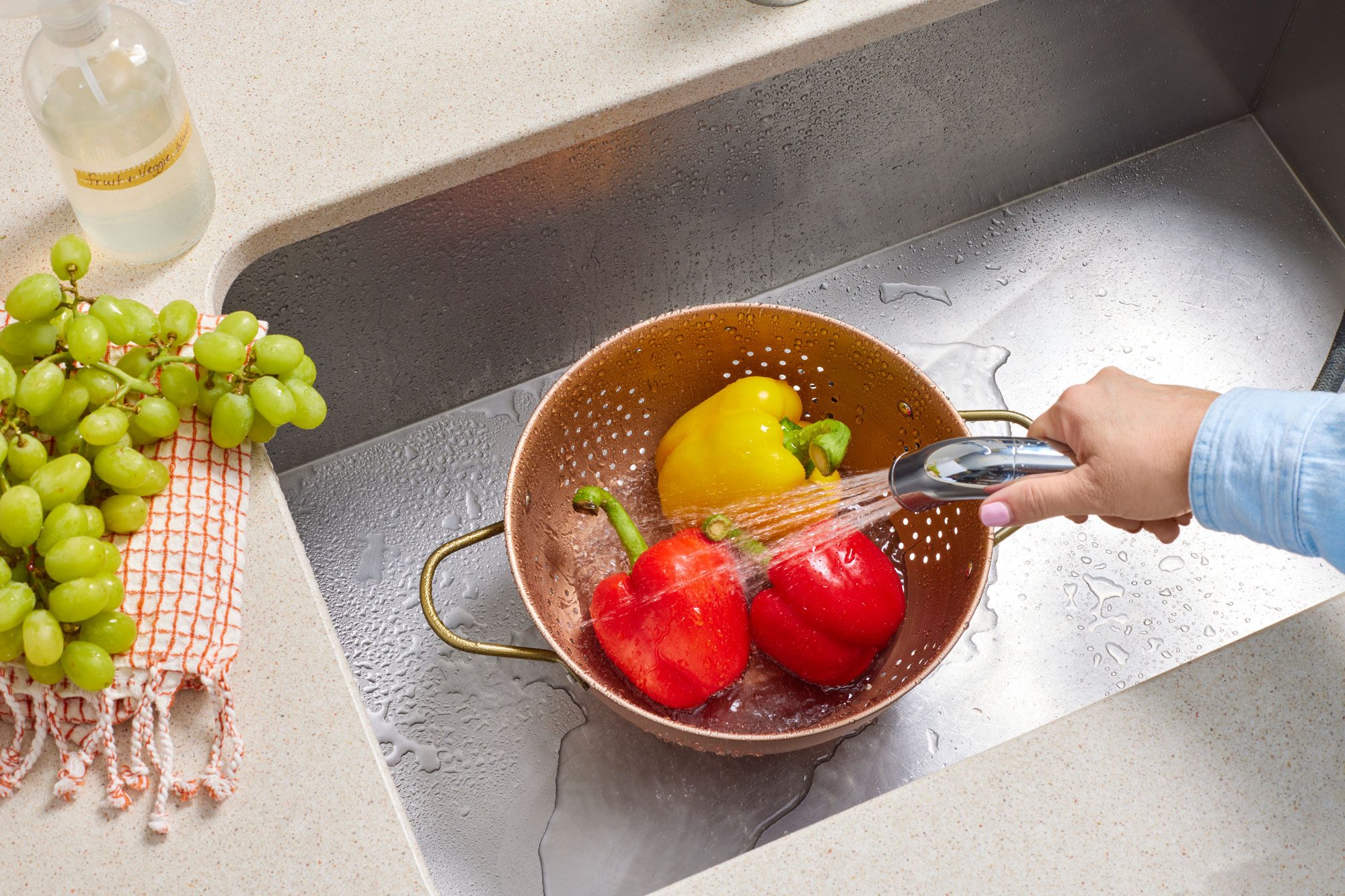Three peppers are in a colander in a stainless steel sink. A hand uses the spray faucet to rinse them.