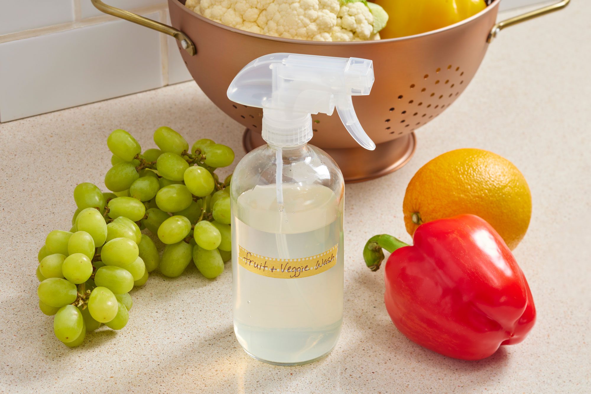 homemade fruit and vegetable wash spray on a kitchen counter with fresh fruit and vegetables arranged on either side of the spray bottle