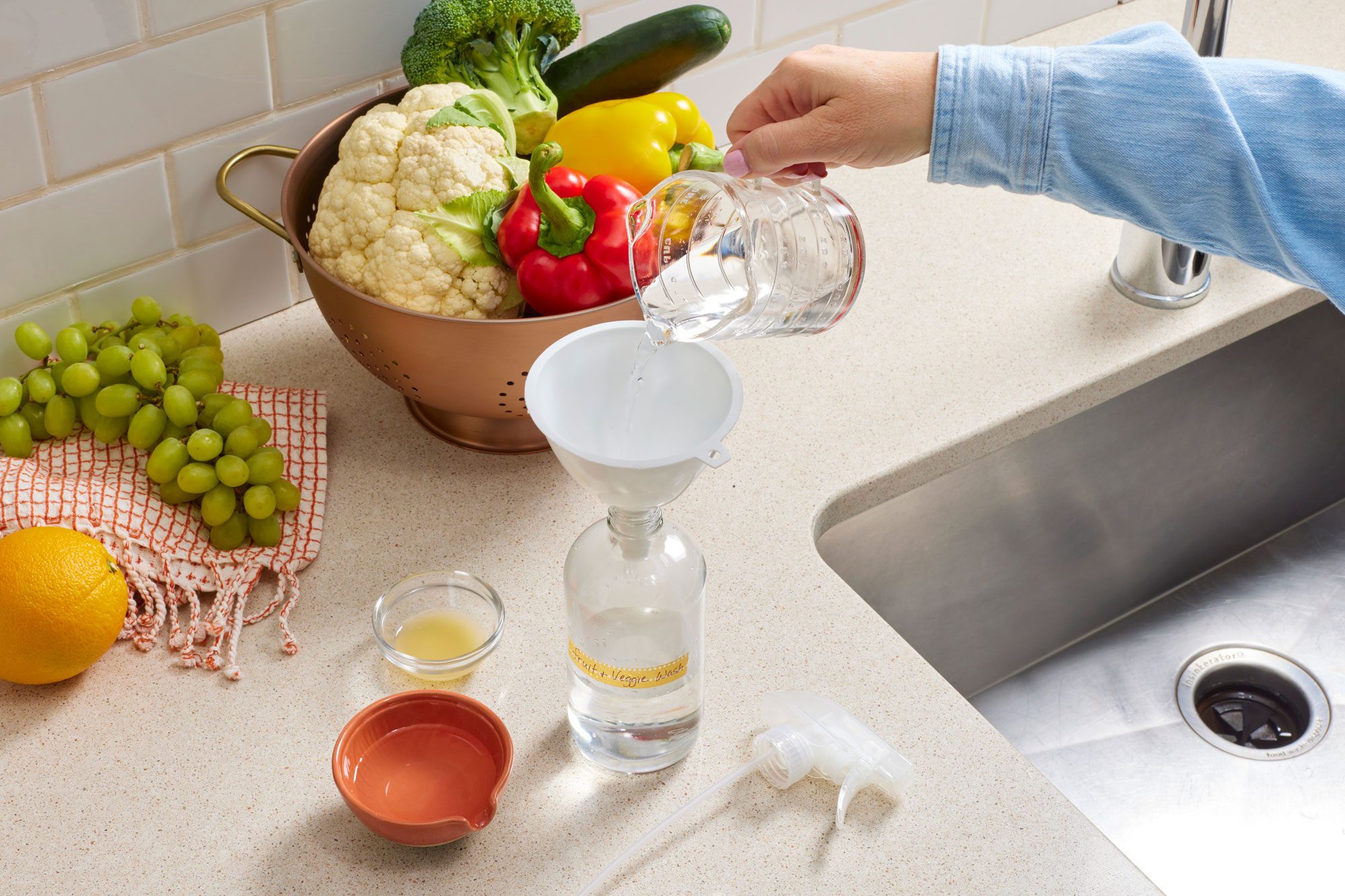 in a kitchen environment near the skin, a hand is pouring homemade fruit and veggie wash ingredients into a spray bottle using a funnel. some fresh fruit and vegetables are on the counter nearby