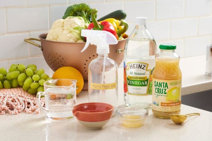 Homemade fruit and vegetable wash supplies arranged on a kitchen countertop near a sink. fresh fruit and veggies are in a colander in the background