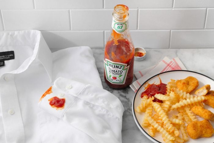 White Shirt Stained With Ketchup next to open bottle of ketchup and french fries and chicken nuggets on a plate; all on marble kitchen counter top