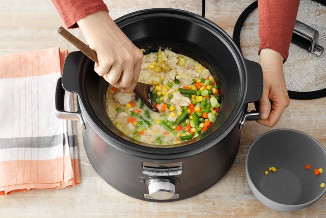 Cooked chicken and vegetables in slow cooker with someone stirring in additional vegetables in the pot.
