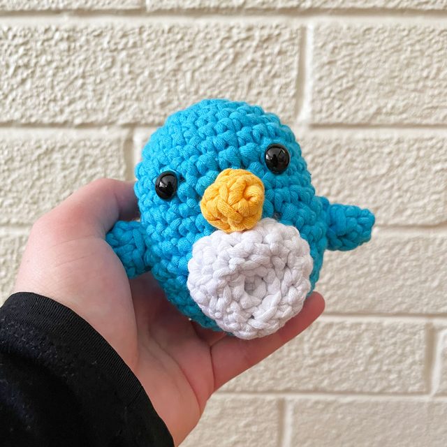 Woobles Crochet Kit finished product Pierre the Penguin being held in a person's hand with a white brick wall background