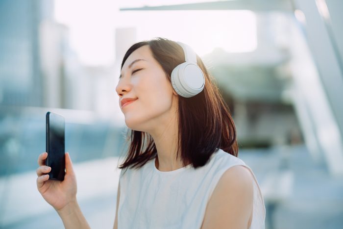 Beautiful young Asian woman with her eyes closed, enjoying music over headphones from her smartphone out in the city, against urban city scene. Music, lifestyle and technology