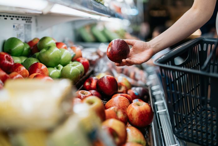 Cropped shot of young Asian woman choosing fresh organic fruits in supermarket. She is picking a red apple along the produce aisle. Routine grocery shopping. Healthy living and eating lifestyle