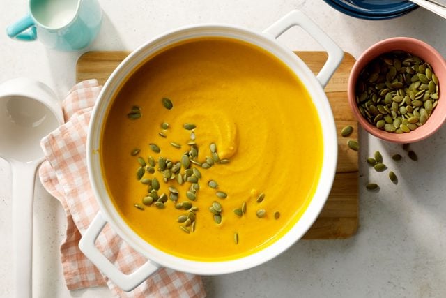 Copycat Panera Autumn Squash Soup in a white cast iron pot sitting on a cutting board with a ladle, pepitas, and cream nearby