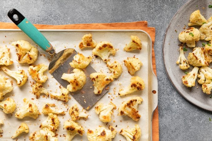 Crispy Cauliflower on a baking sheet with a spatula; some crispy cauliflower pieces are on a plate nearby