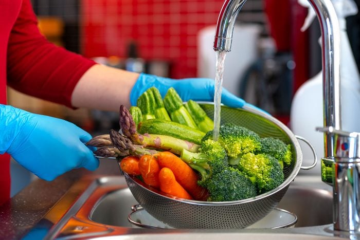 Woman Washes Vegetables in a strainer in the kitchen sink while wearing rubber gloves