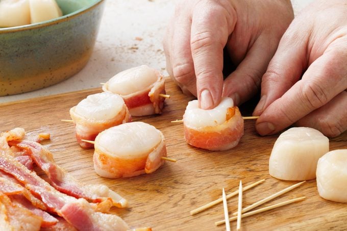 hand wrapping strips of bacon around scallops and securing with a toothpick to prep Bacon Wrapped Scallops