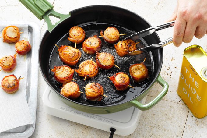 Bacon Wrapped Scallops cooking in a green skillet; hand with tongs is flipping one