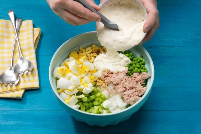 hand pouring the dressing into a bowl containing the other ingredients To Make Tuna Macaroni Salad