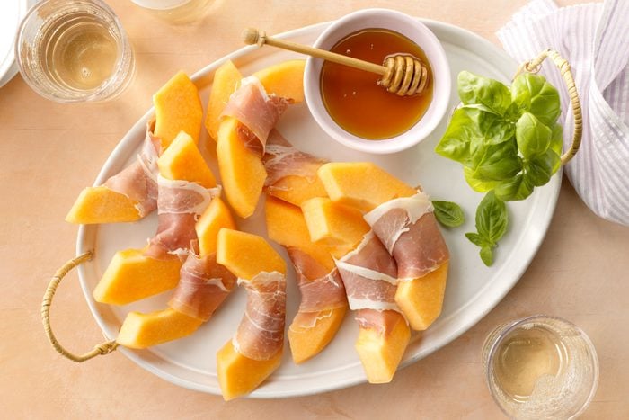 Prosciutto And Melon Appetizer arranged on a tray with honey and basil; on peach surface