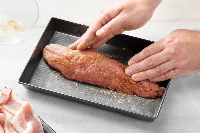 hands adding spice rub to pork tenderloin for Bacon Wrapped Pork Tenderloin