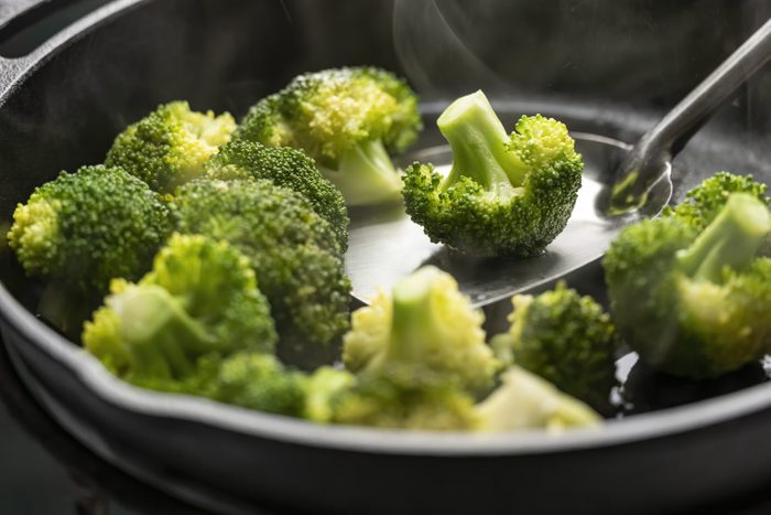 Fresh broccoli spears being sauteed/stir fried in a heavy iron pan on a gas stove against a turquoise colored background.