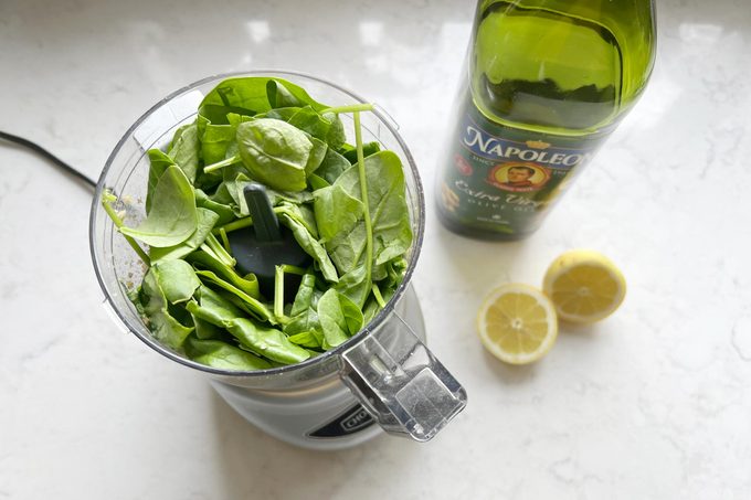 spinach in a food processor with a bottle of olive oil and a sliced lemon off to the side