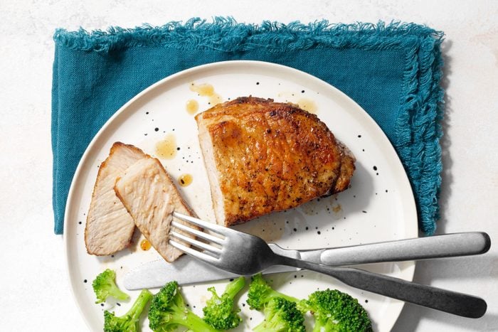 Pork Chop on a plate with a few slices cut; fork, knife, and broccoli also on plate; blue cloth napkin nearby