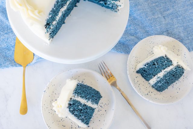 two blue velvet cake slice on a plate and the whole cake on a cake stand in background