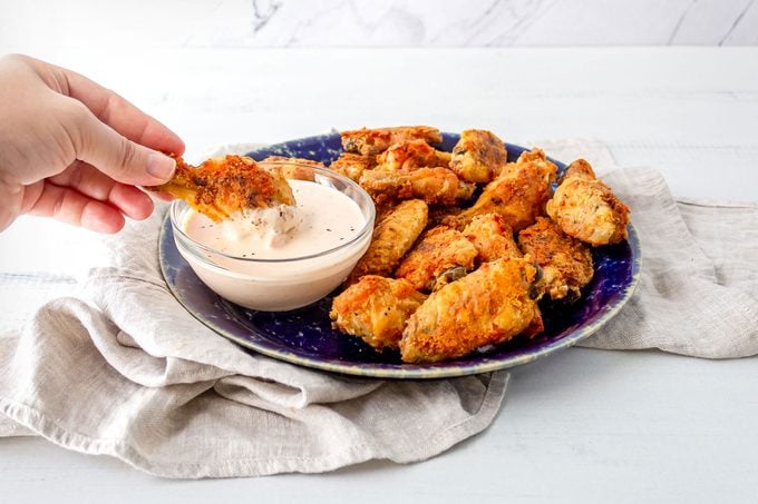 Crispy Oven Fried Chicken Wings Served in Fancy Plate with the Bowl of Spicy Dipping Sauce on Wooden Surface and a hand dipping one piece in sauce