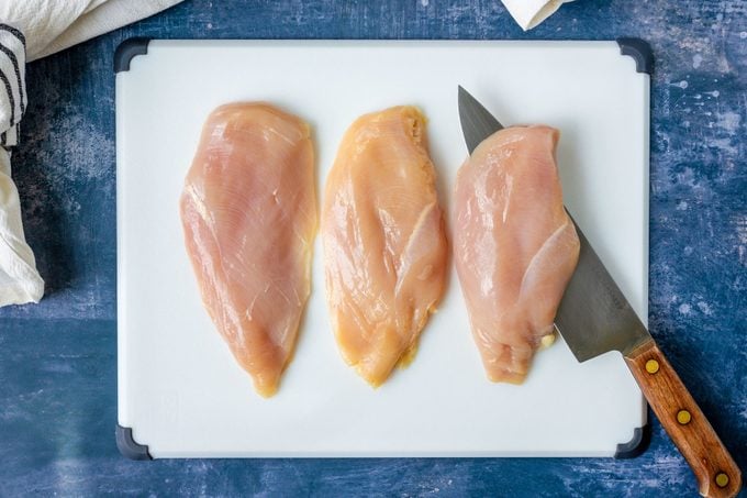 Raw chicken breasts, one with a knife slicing through on white cutting board on a blue surface