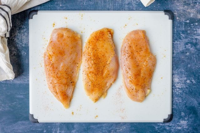 Three chicken breast cutlets with seasoning on white cutting board on blue surface