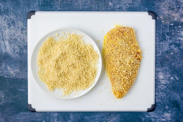 Breaded chicken breast next to plate of breadcrumbs on white cutting board on blue surface