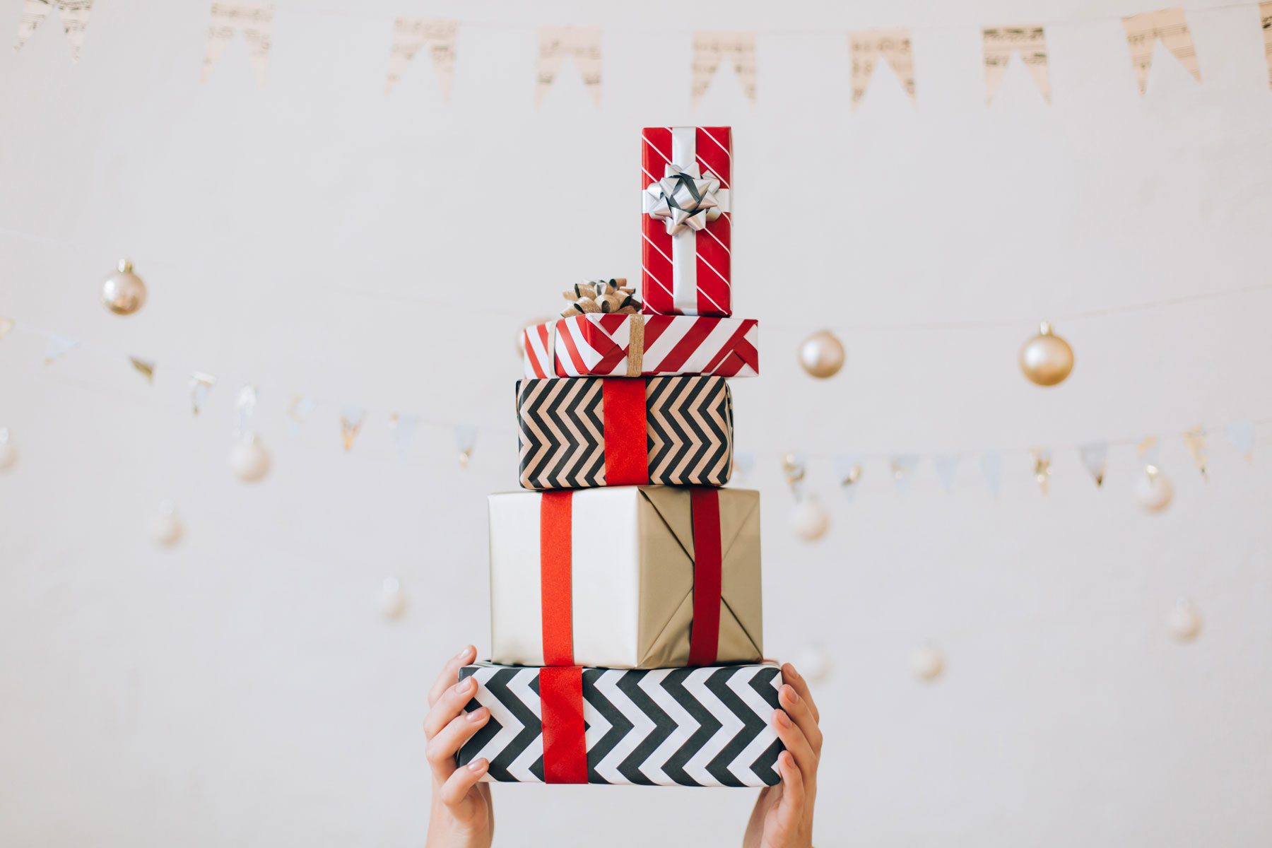 anonymous Hands Are Holding up a Stack Of Gift Boxes With Bows Against White Wall With Garlands On Strings