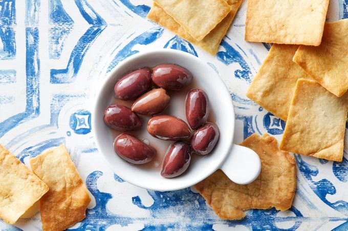 Kalamata olives in a dish on blue and white background with pita chips scattered